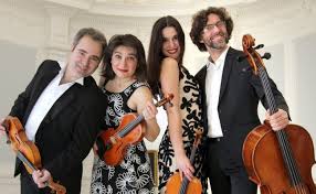 Four musicians in formal attire pose with string instruments—a cello, two violins, and a viola—smiling in an elegant, bright setting reminiscent of St. Nicholas’ Collegiate Church’s ornate white decor.
