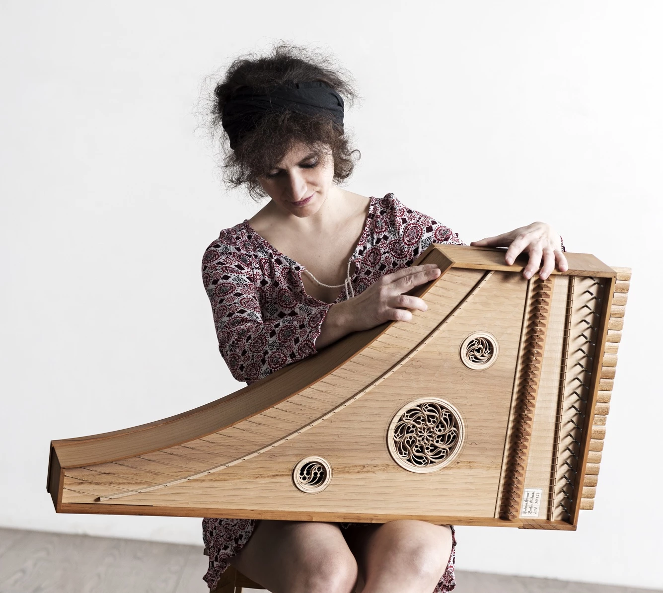 A woman with curly hair and a headband plays a wooden string instrument with intricate carvings, seated on a stool against a plain, light background—reminiscent of performances at St. Nicholas’ Collegiate Church.
