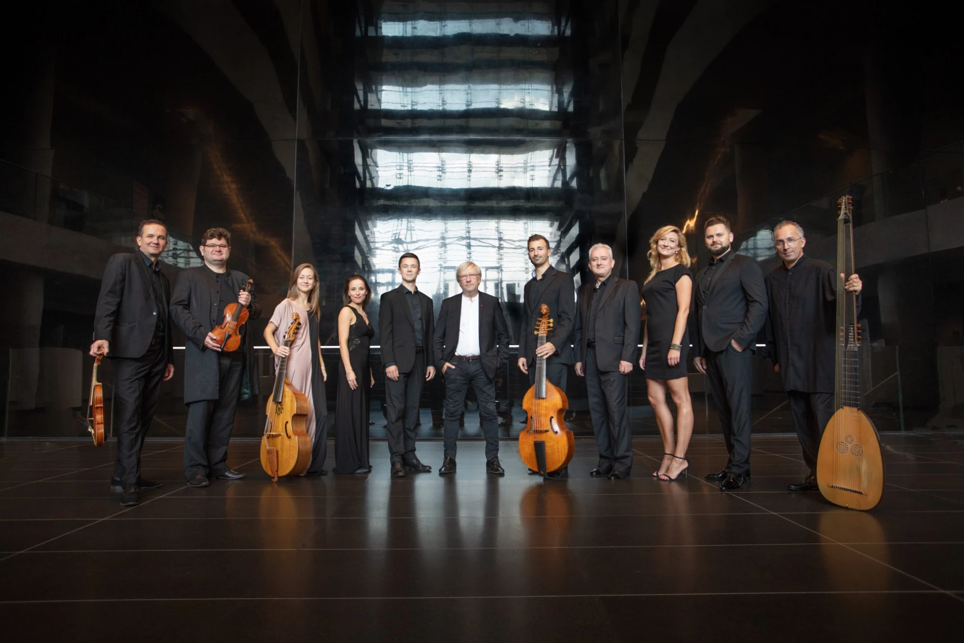 A group of twelve musicians, dressed in formal black attire, stand in a modern, reflective hall reminiscent of St. Nicholas’ Collegiate Church. Several hold string instruments as they line up, facing the camera and smiling warmly.