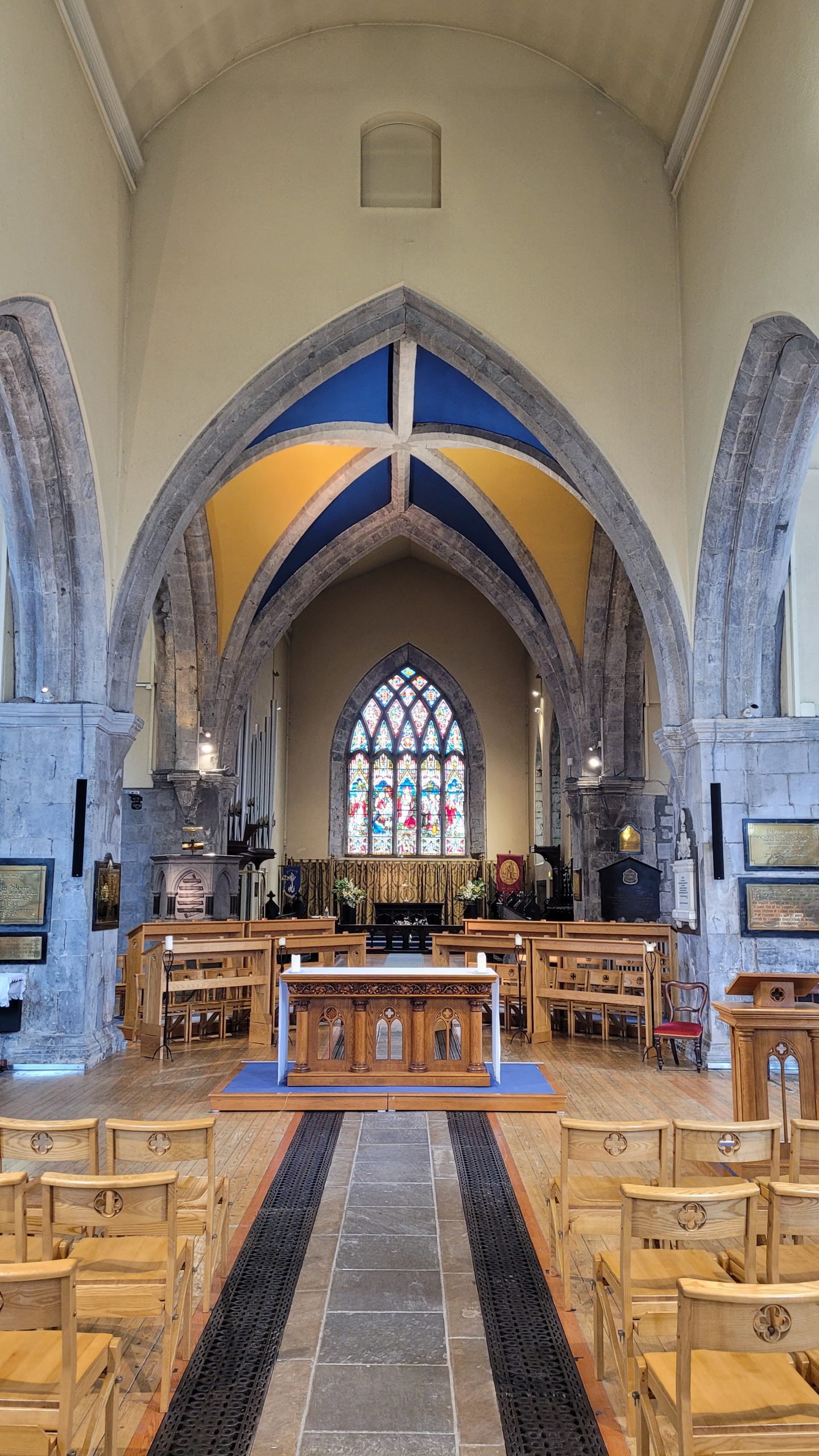 View of St. Nicholas’ Collegiate Church interior with wooden chairs, a central aisle, ornate altar, pointed stone arches, and a colorful stained glass window at the far end. The vaulted ceiling displays blue and yellow sections.