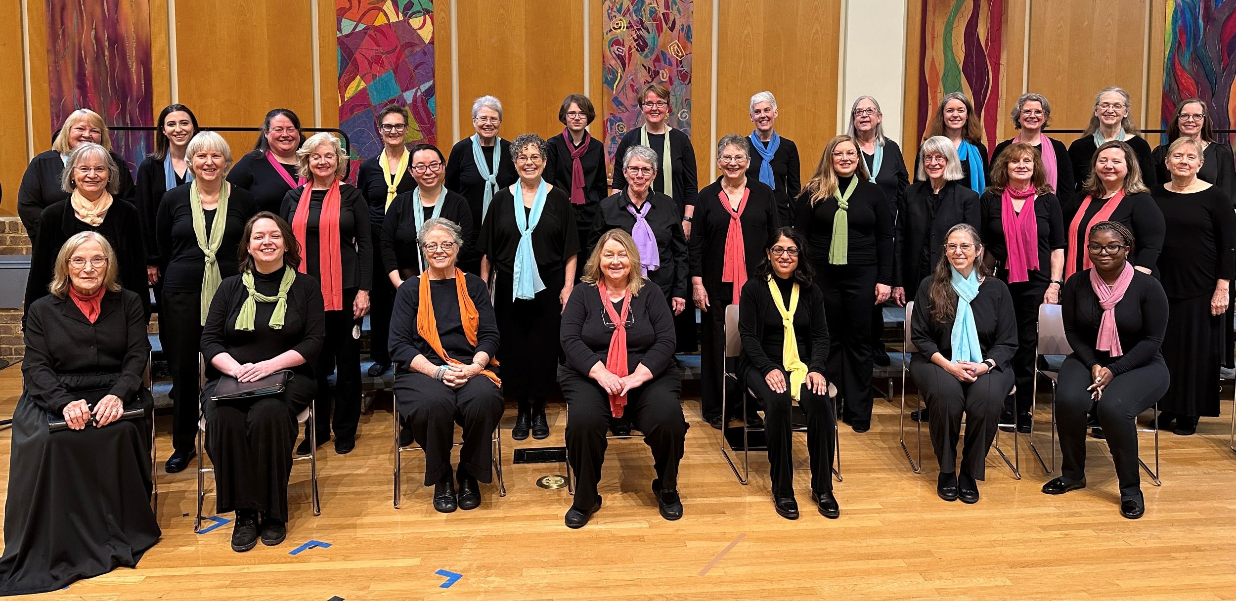 A group of women dressed in black with colorful scarves pose for a choir photo indoors at St. Nicholas’ Collegiate Church, standing and sitting in rows on a wooden floor with vibrant abstract art panels in the background.