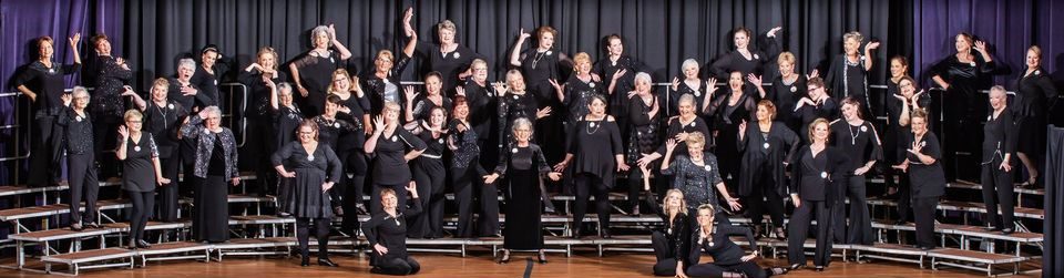A large group of women dressed in black pose joyfully on choir risers in St. Nicholas’ Collegiate Church, many with one hand raised and smiling, suggesting a celebratory or theatrical choir performance before a dark curtain.