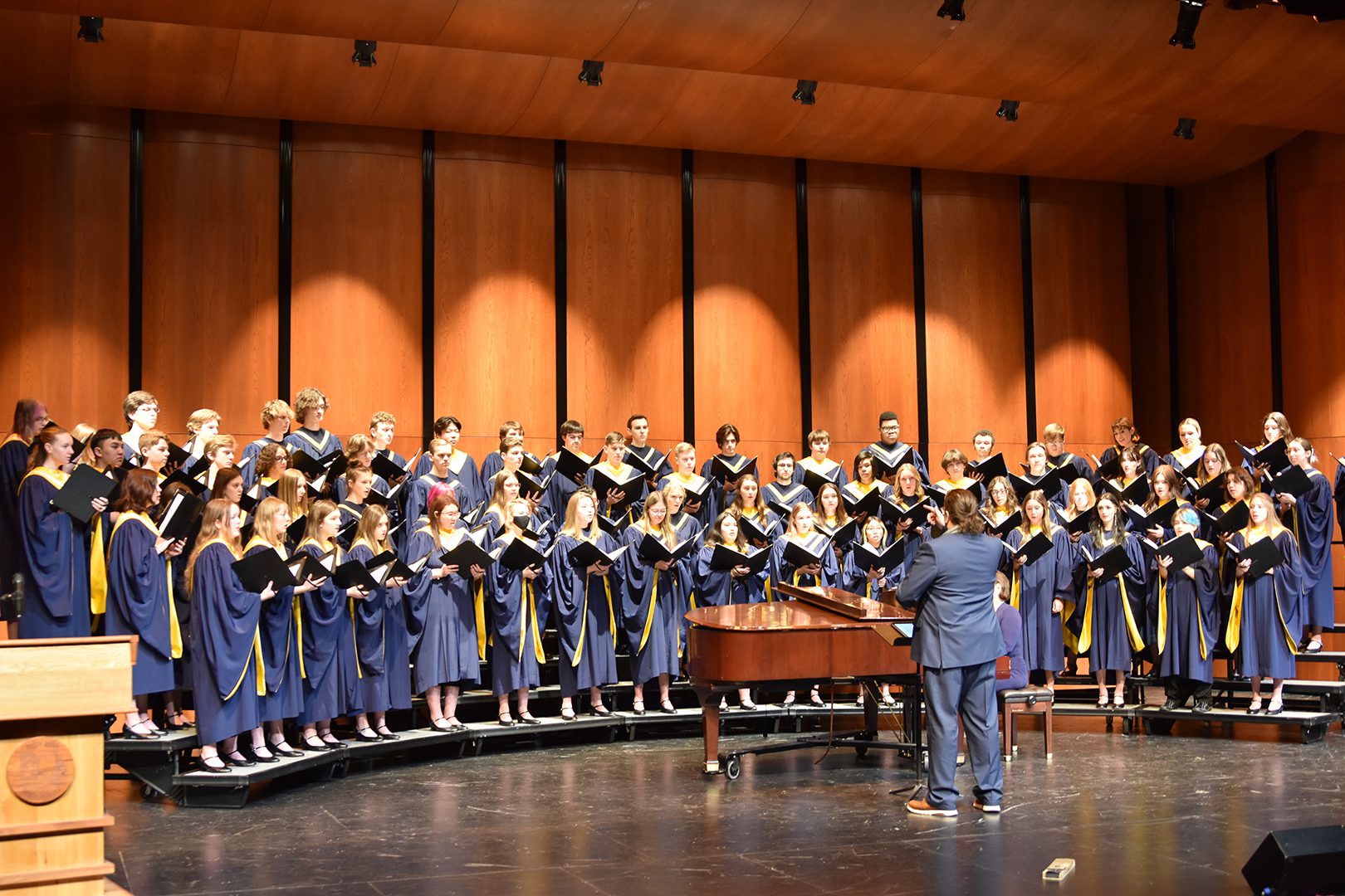 The Prior Lake Choir, dressed in navy blue robes, sings on stage behind a grand piano, standing on risers as a conductor in a suit leads them in a wood-paneled concert hall.