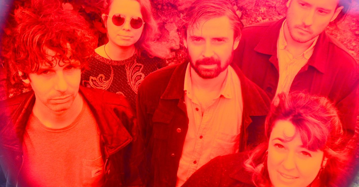 Five people stand closely together, looking up at the camera with neutral expressions. The image, set near St. Nicholas’ Collegiate Church, has a strong red and pink filter, creating a vibrant, surreal effect.