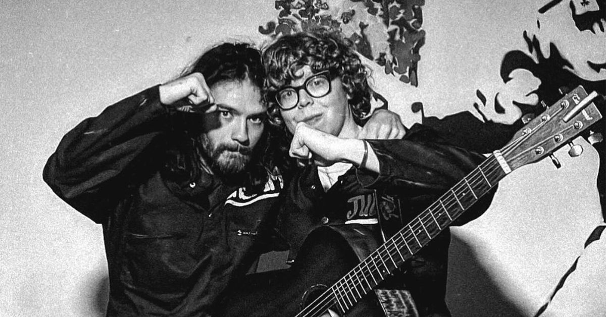 Two young people pose together in black and white, each raising a fist to their chin. One has long hair and a beard, while the other wears glasses and holds an acoustic guitar. Behind them, abstract art hints at St. Nicholas’ Collegiate Church nearby.