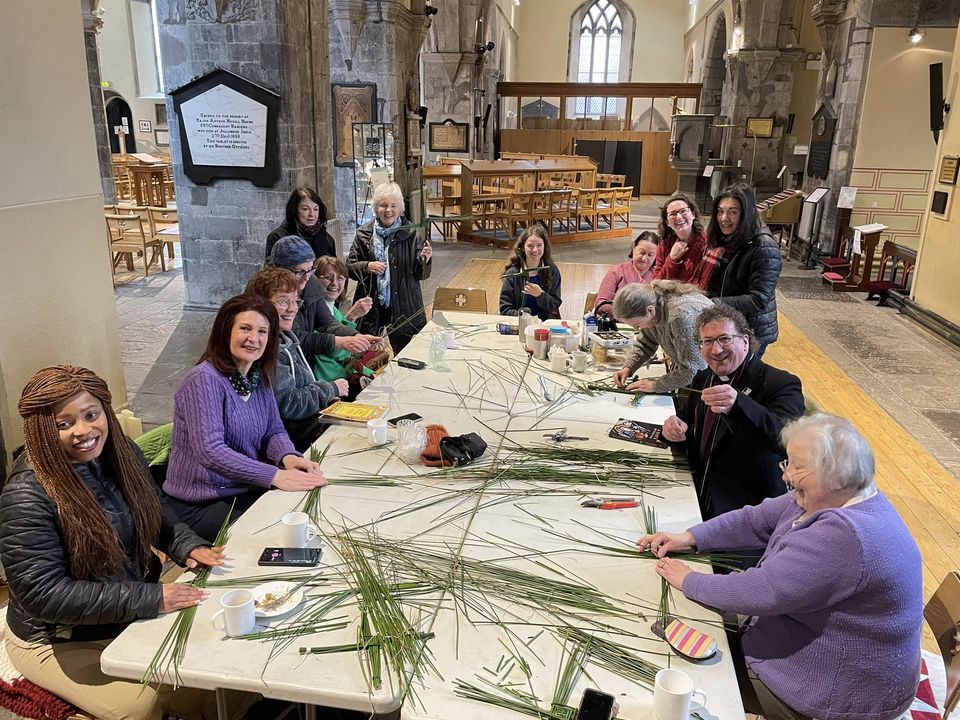 A group of people sit around tables inside St. Nicholas’ Collegiate Church, weaving long palm leaves. Some smile at the camera while others focus on their work. Cups and books are scattered on tables, with church pews visible in the background.