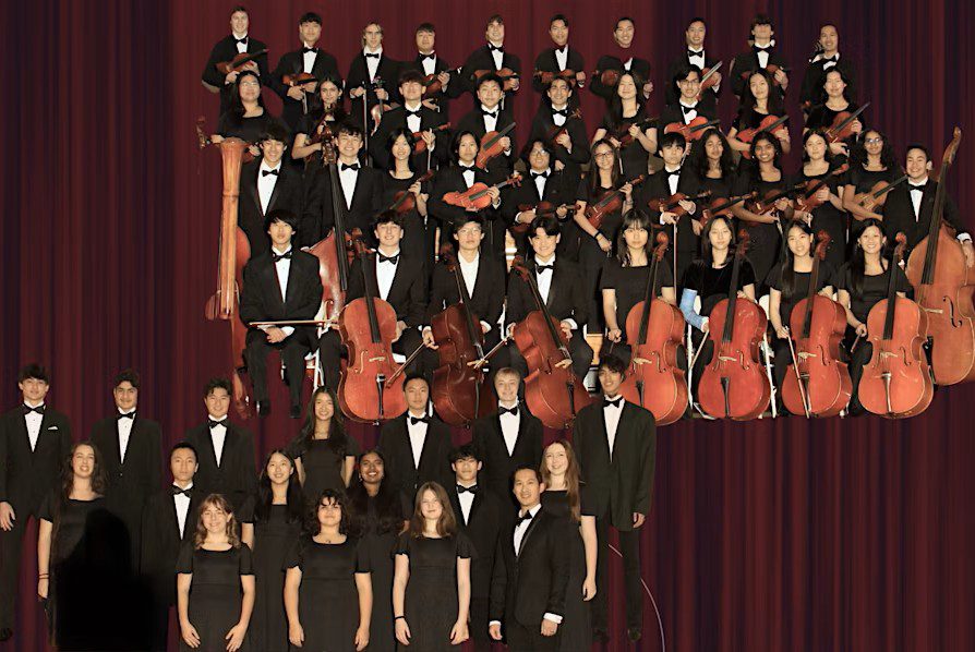 A large orchestra group poses on stage in formal black attire with string instruments such as violins, violas, and cellos, arranged in several rows before a maroon curtain at St. Nicholas’ Collegiate Church.