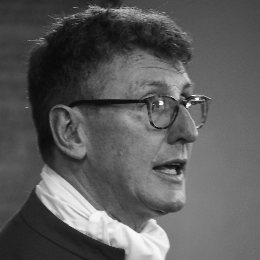 Black and white photo of an older man with short curly hair and glasses, wearing a suit and a cravat, speaking or mid-sentence, shown in profile with a blurred background.