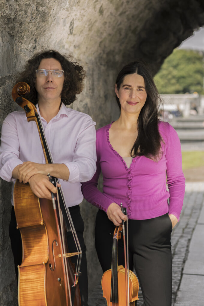 A man with curly hair and glasses holds a cello, while a woman in a pink cardigan holds a violin. Members of ConTempo, they stand side by side under a stone archway outdoors, smiling as if ready for an inspiring lunchtime performance.