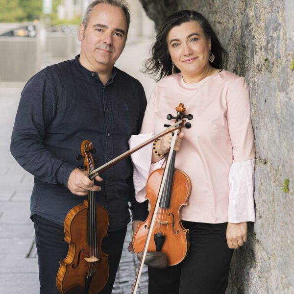 A man and a woman stand side by side outdoors, each holding a violin. The woman is leaning against a stone wall, and both are smiling at the camera—ready to perform at a ConTempo Lunchtime Concert.