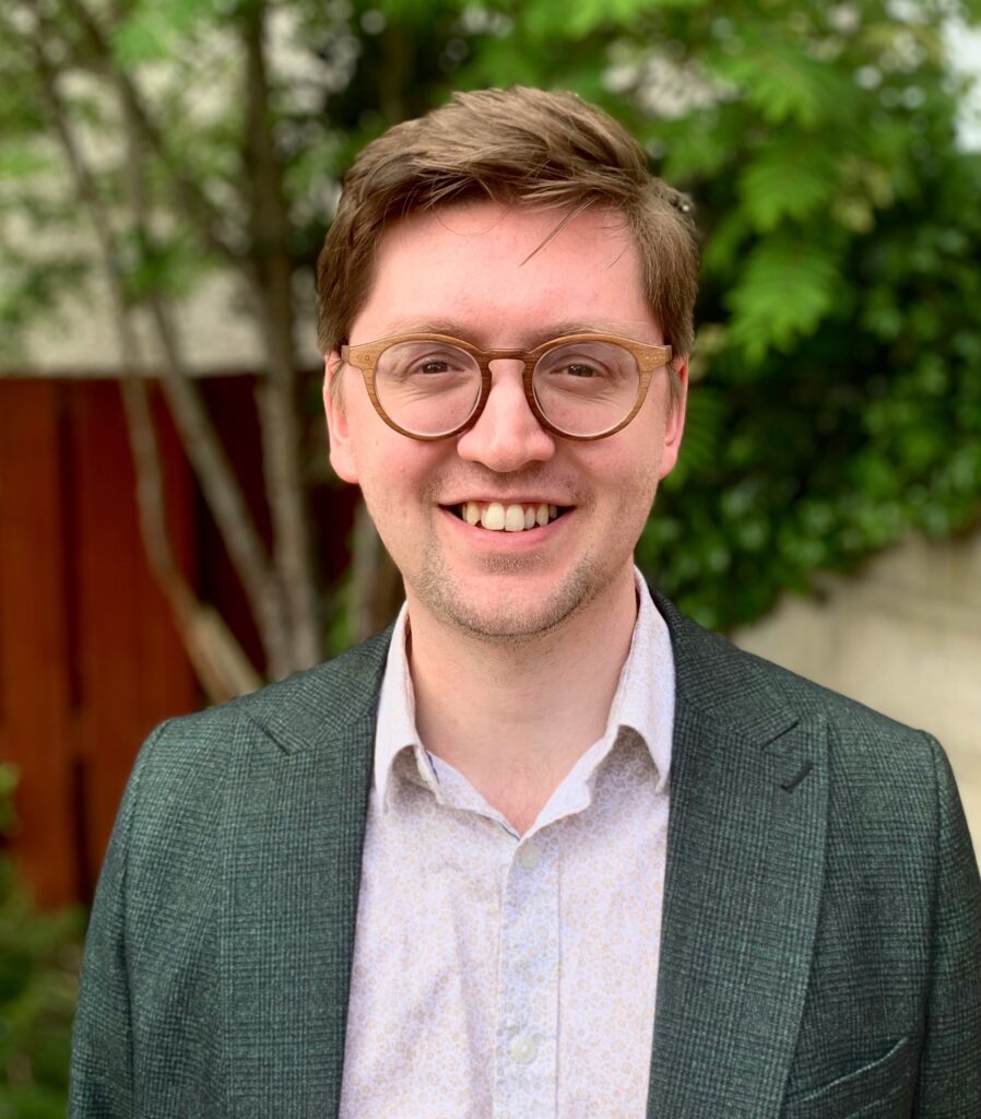 A young man with short brown hair and glasses smiles at the camera. He is wearing a light-colored shirt and a green blazer, standing outdoors with greenery and a wooden fence in the background.