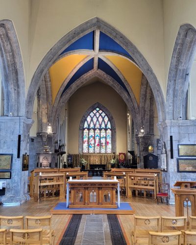 View of St. Nicholas’ Collegiate Church interior with wooden chairs, a central aisle, ornate altar, pointed stone arches, and a colorful stained glass window at the far end. The vaulted ceiling displays blue and yellow sections.