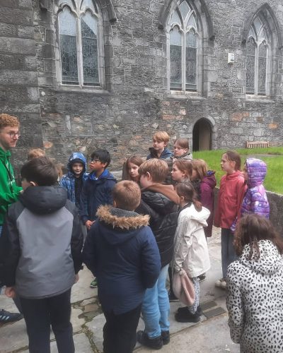 A group of children wearing jackets stands outside St. Nicholas’ Collegiate Church, a stone building with arched windows, listening to an adult guide in a green hoodie. The scene appears to be an educational or historic tour.