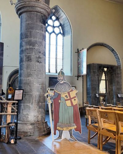 A cardboard cutout of a medieval knight in armor stands beside a stone pillar inside St. Nicholas’ Collegiate Church, surrounded by arched windows and wooden chairs.