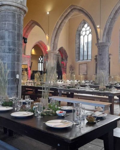 Elegant dining tables set with plates, glasses, and decorative grass centerpieces are arranged in rows inside the grand hall of St. Nicholas’ Collegiate Church, featuring stone columns, arched windows, and high vaulted ceilings.