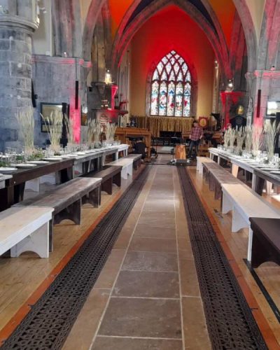 Long wooden tables with benches are set for a meal inside St. Nicholas’ Collegiate Church, with high arched ceilings, stained glass windows, and red lighting highlighting the stone walls and altar area.