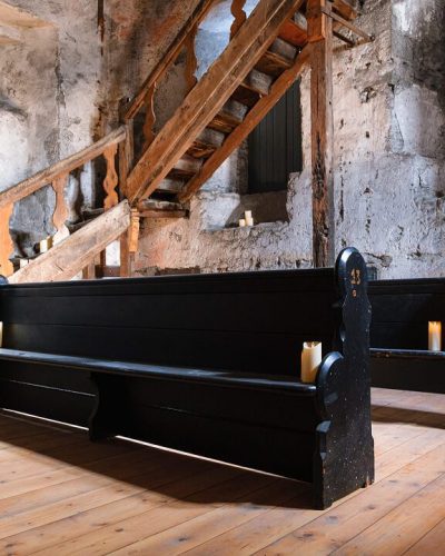 Two black wooden pews sit on a polished wooden floor in a rustic, stone-walled room at St. Nicholas’ Collegiate Church. A wooden staircase with a carved railing ascends at the back, while candles placed around the room create a warm atmosphere.